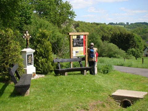 Infotafel am Böllweg Zwei Personen lesen eine Infotafel in einer grünen, hügeligen Landschaft mit Bäumen und Bänken.