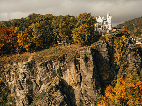 Schäferwand Ein weißes Schloss mit Türmen steht auf einer steilen Klippe, umgeben von herbstlich gefärbten Bäumen unter einem bewölkten Himmel.