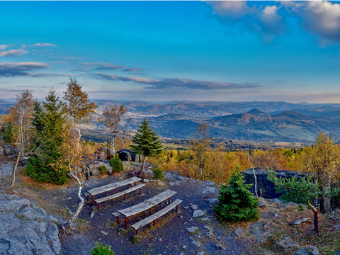 Zwei Holzbänke auf einem felsigen Aussichtspunkt mit Blick auf bewaldete Hügel und ein Tal unter einem klaren blauen Himmel.