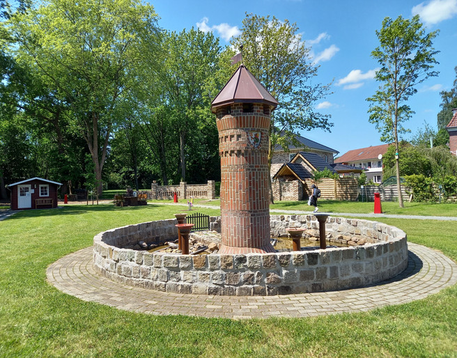 Mehrgenerationenplatz "Lüttje Festung" in Apen Der Mehrgenerationenplatz "Lüttje Festung" in Apen zeigt einen zentralen Backsteinturm inmitten einer kreisförmigen Steinmauer, umgeben von gepflegtem Rasen und Bäumen.
