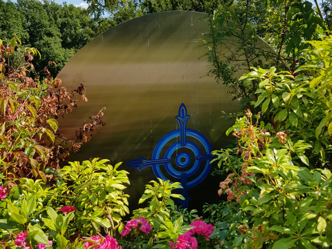 Stockhausen Grabstätte Rückseite Eine metallene Skulptur mit blauem Kompassmotiv, umgeben von üppigem Grün und blühenden Blumen.