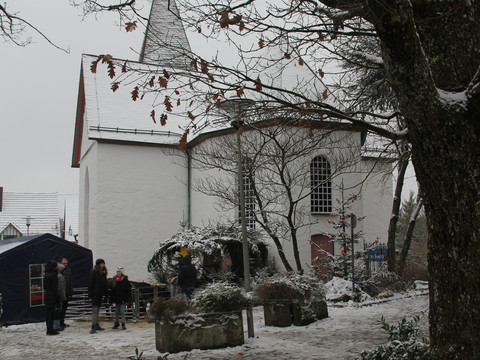 Bonte Kerke Lieberhausen Eine verschneite Kirche mit steilem Dach, umrahmt von Bäumen und wenigen Personen im Vorgarten.