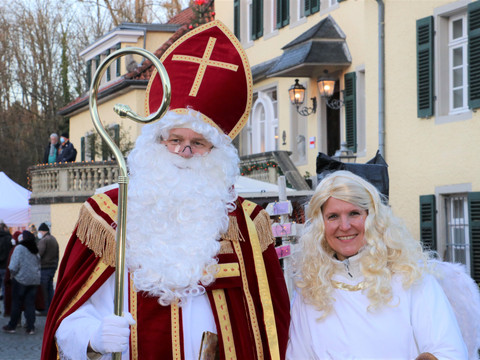 Weihnachtsmarkt Schloss Eulenbroich Sankt Nikolaus mit Bischofsstab und Engel auf einem festlichen Weihnachtsmarkt, im Hintergrund Fachwerkhäuser.