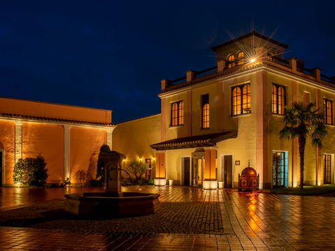 Mediterana Ein beleuchtetes Gebäude mit mediterranem Stil bei Nacht, umgeben von einem gepflasterten Hof.