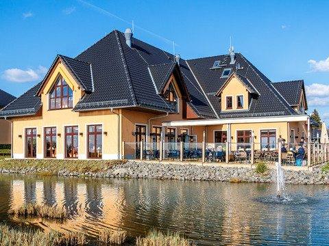 Restauracja Seeperle © Seepark Auenhain.jpg Restauracja z tarasem, położona nad małym stawem, nowoczesna architektura, błękitne niebo.