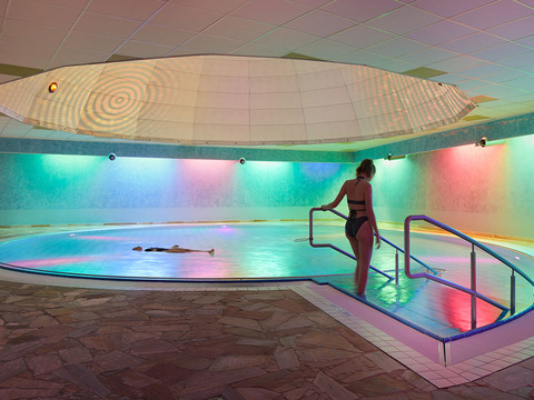 Toskana Therme Bad Schandau: Eine Frau in einem schwarzen Badeanzug betritt ein Hallenbad mit bunten Lichtern; eine Person schwimmt im Wasser.