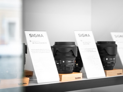 SIGMA Objektive in derAusleihe in Bad Schandau Drei Sigma-Kameraobjektive auf Holzständern in einem Regal, begleitet von Informationskarten. Die Atmosphäre ist modern und minimalistisch.