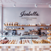 Isabella Glutenfreie Patisserie Blick auf eine Theke, hinter deren Glas kleine Gepäckstücke mit Beeren sowie Brotwaren ausgelegt sind. Auf der Wand dahinter steht "Isabella Glutenfreie Patisserie seit 2014" in Großbuchstaben. Darunter ist ein langes Holzbrett an der Wand befestigt, auf dem Gläser, Tassen, Gewürze sowie eine Topfpflanze stehen. Auf zwei kleineren Holzbrettern darunter stehen weitere Tassen und gestapelte Untertassen. Auf dem Küchentisch darunter steht eine große Kaffeemaschine sowie weitere Küchenutensilien.