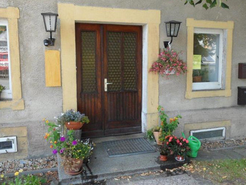 Hofladen Ziegenhof Lauterbach Holztür mit gelbem Rahmen, flankiert von Fenstern und Laternen; bunte Blumentöpfe und grüne Gießkanne auf dem Boden, einladende Atmosphäre.