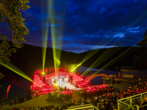 Evening stage performance with lighting effects, illuminated lake and audience in an open-air setting.