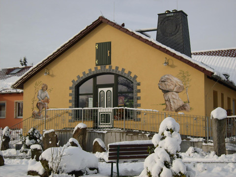Gold- und Mineralien Erlebnisstätte Hohwald  Gelbes Gebäude mit bemalter Fassade, zeigt eine Person und Felsen, umgeben von schneebedeckten Büschen und Steinen, winterliche Atmosphäre.
