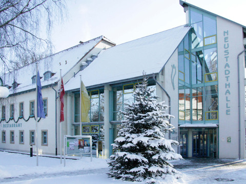 Verschneite Neustadthalle mit Glasfassade, flankiert von Fahnen und einem schneebedeckten Tannenbaum, bei klarem Himmel.