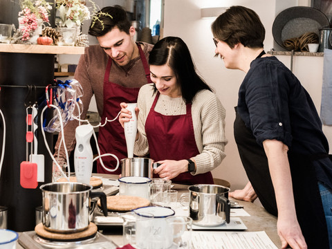 Seifen selber machen  Drei Personen in roten Schürzen kochen gemeinsam in einer Küche, verwenden einen Stabmixer und wirken konzentriert und fröhlich.