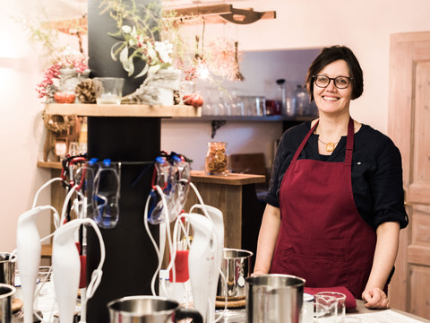 Seifenschneiderin Eine lächelnde Frau in roter Schürze steht in einer Küche mit Mixern und Dekorationen aus Pflanzen und Gläsern im Hintergrund.