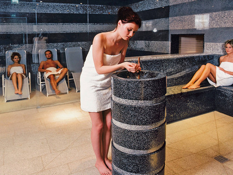 Sauna im Geibeltbad Pirna: Eine Frau im weißen Handtuch wäscht ihre Hände an einem grauen Brunnen in einem Spa, während zwei Personen im Hintergrund auf Liegen entspannen.