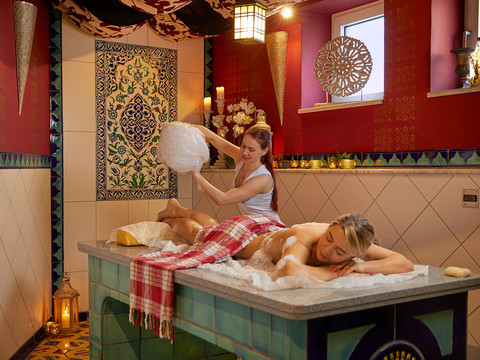 Eine Frau erhält eine Schaummassage auf einem Marmor-Tisch in einem orientalisch dekorierten Spa mit roten Wänden und Kerzenlicht.