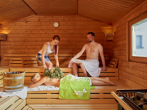 Drei Personen in einer Holzsauna; eine Frau liegt auf einer Bank mit einem grünen Handtuch, während eine andere Frau sie mit Zweigen massiert. Ein Mann sitzt daneben.