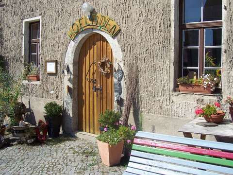 Hofladen Gut Gamig Eingang eines Hofladens mit Holztür, umgeben von Pflanzen in Töpfen; daneben bunte Bank und Tisch mit Blumentöpfen, sonnige Atmosphäre.