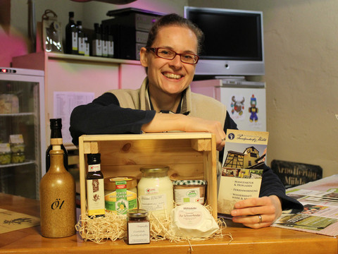 Eine lächelnde Person präsentiert einen Geschenkkorb mit regionalen Produkten wie Öl, Honig und Marmelade auf einem Holztisch in einem gemütlichen Laden.