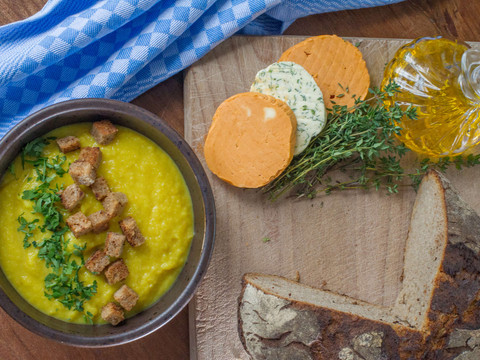 Mühlenhof bar Schmilka Gelbe Suppe mit Croutons und Petersilie in einer Schüssel, daneben Brot, Kräuterbutter und Olivenöl auf einem Holzbrett, mit blau-weißem Tuch.