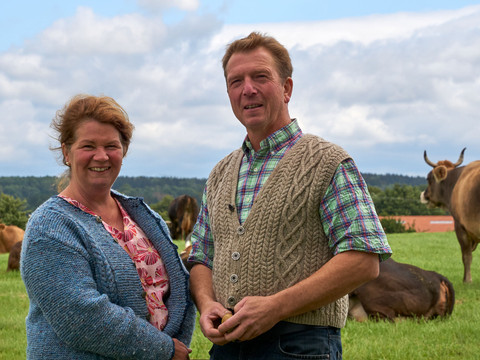 Ein Mann und eine Frau stehen lächelnd auf einer grünen Wiese mit Kühen im Hintergrund, unter einem bewölkten Himmel.