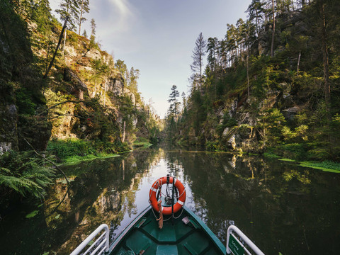Ein grünes Boot mit Rettungsring fährt durch eine schmale, von Bäumen und Felsen gesäumte Schlucht, die sich im ruhigen Wasser spiegelt.