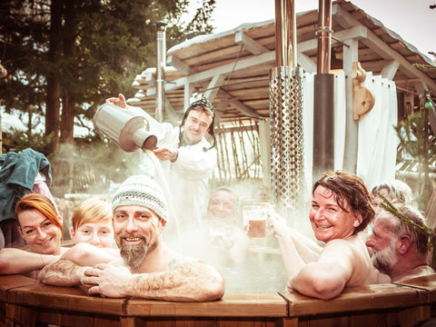 Badezuberei Schmilka Gruppe von Menschen entspannt in einem dampfenden Holzbottich im Freien, während eine Person heißes Wasser nachgießt; fröhliche Stimmung.