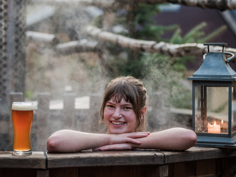 Badezuberei Winterdorf Schmilka Eine Frau lächelt in einem dampfenden Holzbottich, daneben ein Glas Bier und eine brennende Kerze in einer Laterne.