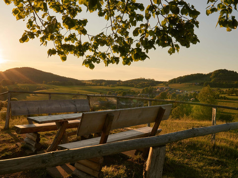 Goßdorfer Hankehübel Aussicht Holzbänke und Tisch auf einem Hügel mit Blick auf eine grüne Landschaft bei Sonnenuntergang, umrahmt von Baumzweigen.