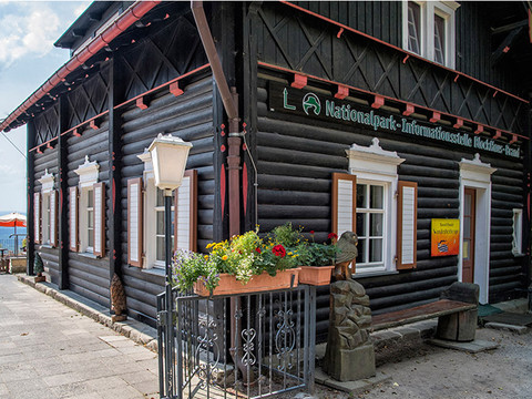 Informační místo národního parku na značce Schwarzes Holzhaus der Nationalpark-Informationsstelle mit Blumenkasten und Laterne, daneben eine Bank und ein Sonnenschirm im Hintergrund.