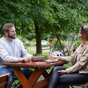 Zwei Personen sitzen an einem Holztisch im Freien; im Hintergrund grüner Garten mit Spielgeräten.