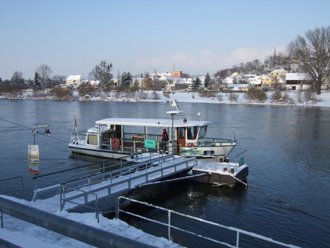 Ein kleines Fährschiff liegt an einem verschneiten Ufer, im Hintergrund sind schneebedeckte Häuser und Bäume zu sehen.