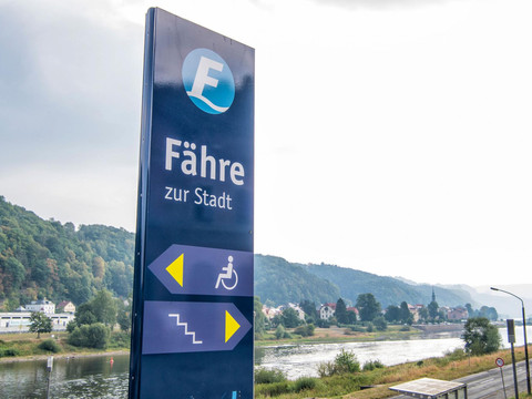 Blaues Schild mit der Aufschrift "Fähre zur Stadt" und Piktogrammen für Rollstuhlzugang und Treppen, im Hintergrund Fluss und bewaldete Hügel.