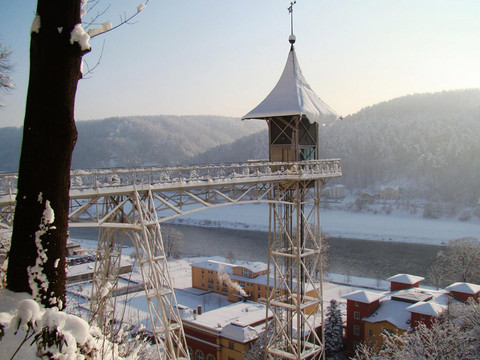Historischer Personenaufzug Bad Schandau im Winter Ein schneebedeckter Aussichtsturm mit spitzem Dach steht über einem Fluss, umgeben von verschneiten Hügeln und bunten Gebäuden.A snow-covered observation tower with a pointed roof stands above a river, surrounded by snow-covered hills and colorful buildings.Nad řekou se tyčí zasněžená rozhledna se špičatou střechou, obklopená zasněženými kopci a barevnými budovami.Pokryta śniegiem wieża obserwacyjna ze spiczastym dachem stoi nad rzeką, otoczona ośnieżonymi wzgórzami i kolorowymi budynkami.Een besneeuwde uitkijktoren met een puntdak staat boven een rivier, omringd door besneeuwde heuvels en kleurrijke gebouwen.Una torre di osservazione innevata con un tetto a punta si erge sopra un fiume, circondata da colline innevate ed edifici colorati.
