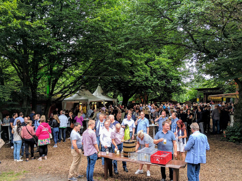 Eine Menschenmenge genießt an einem sonnigen Tag ein geselliges Event im Freien, umgeben von Bäumen.