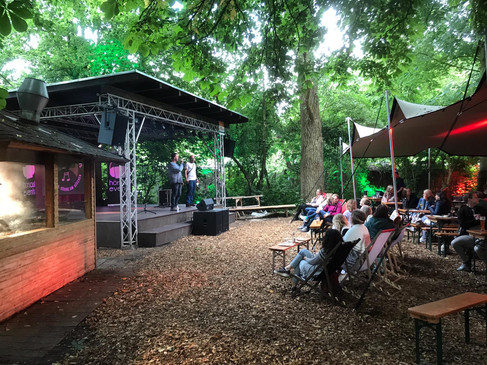 Eine Open-Air-Bühne mit Zuschauern, umgeben von üppigen Bäumen in einem stimmungsvollen Setting.