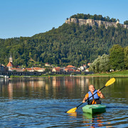 Kanu Aktiv Tours Ein Mann paddelt in einem grünen Kajak auf einem Fluss, im Hintergrund eine Stadt mit Kirche und bewaldetem Hügel unter klarem Himmel.