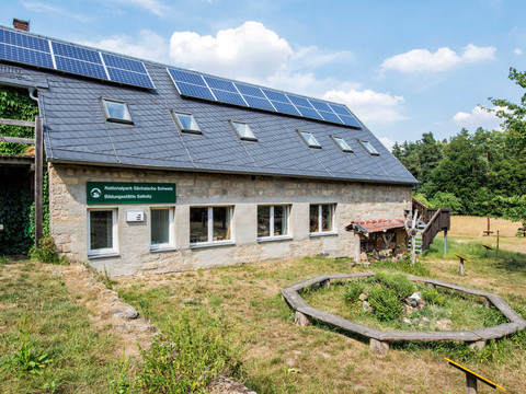 Steinhaus mit Solarpanels auf dem Dach, umgeben von grüner Wiese und Bäumen, mit einem Schild "Nationalpark Sächsische Schweiz Bildungsstätte Sellnitz".