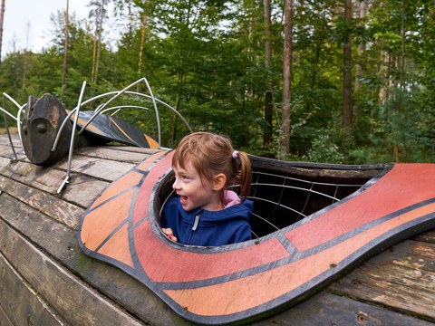 Ein lachendes Kind in blauer Jacke schaut aus einem hölzernen Spielgerät, das wie ein Insekt gestaltet ist, in einem Wald.A laughing child in a blue jacket looks out of a wooden play structure designed like an insect in a forest.Smějící se dítě v modré bundě vyhlíží z dřevěné hrací konstrukce navržené jako hmyz v lese.Roześmiane dziecko w niebieskiej kurtce wygląda z drewnianej konstrukcji do zabawy zaprojektowanej jak owad w lesie.Een lachend kind in een blauwe jas kijkt uit een houten speelstructuur die is ontworpen als een insect in een bos.Un bambino ridente con una giacca blu si affaccia da una struttura di gioco in legno disegnata come un insetto in una foresta.