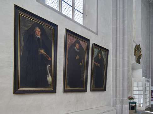 Drei historische Porträtgemälde an einer Kircheninnenwand, sanft beleuchtet, mit gotischem Fenster darüber.