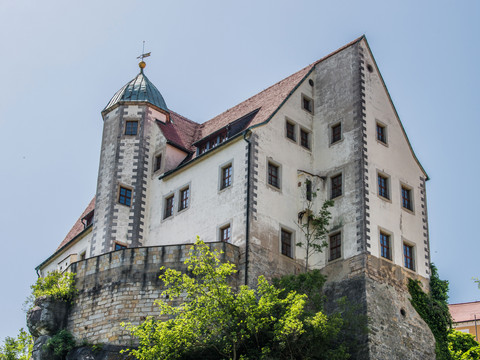 Hrad Hohnstein Historisches Schloss mit weißer Fassade und rotem Ziegeldach auf einem Felsen, umgeben von grünen Bäumen, unter klarem, blauem Himmel.