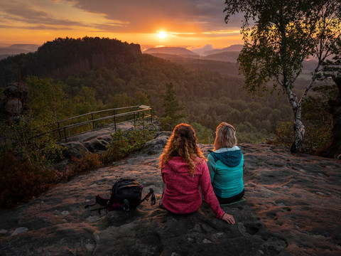 Uitzicht bij zonsondergang vanaf Papststein Zwei Personen sitzen auf einem Felsen und blicken in einen bewaldeten Sonnenuntergang. Eine trägt eine rote, die andere eine blaue Jacke.