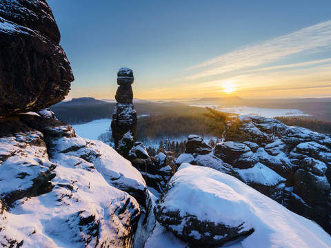 Blick beim Sonnenuntergang vom Pfaffenstein auf die Barbarine Verschneite Felsformationen des Pfaffenstein bei Sonnenuntergang, mit einem markanten, turmartigen Felsen im Vordergrund und einem weiten Tal im Hintergrund.