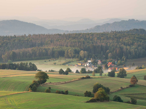 View from the Weifberg Tower Grüne Hügel mit vereinzelten Bäumen, ein kleines Dorf im Tal und neblige Atmosphäre, umgeben von dichten Wäldern und sanften Bergen im Hintergrund.