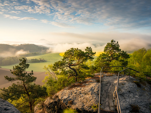 Pohled z císařské koruny Felsvorsprung mit Geländer, umgeben von grünen Bäumen, mit Blick auf neblige Felder und Hügel im Morgenlicht.Rocky outcrop with railings, surrounded by green trees, with a view of misty fields and hills in the morning light.Skalnatý výběžek se zábradlím, obklopený zelenými stromy, s výhledem na mlhavá pole a kopce v ranním světle.Skalista skała z balustradą, otoczona zielonymi drzewami, z widokiem na zamglone pola i wzgórza w porannym świetle.Rotsachtige uitloper met hekwerk, omringd door groene bomen, met uitzicht op mistige velden en heuvels in het ochtendlicht.Un affioramento roccioso con ringhiera, circondato da alberi verdi, con vista su campi e colline nebbiose nella luce del mattino.
