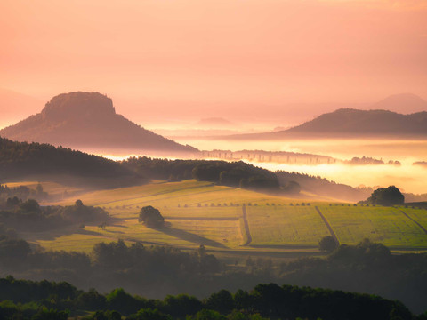 Pohled z vrcholu Kleiner Bärenstein Sanfte Hügel und Felder im Morgennebel, beleuchtet von einem warmen, orangefarbenen Sonnenaufgang, mit einem markanten Berg im Hintergrund.