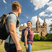 Hoexter-Corvey-Teutoburger-Wald-Tourismus-D-Ketz-074.jpg Ein Paar mit Rucksäcken spaziert lächelnd vor einer historischen Kathedrale mit zwei Türmen.