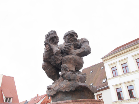 Pomník Ringelnatz ve Wurzenu©SIESING.jpg Bronzová socha zamyšleně sedícího muže s červenými taškovými střechami v pozadí.