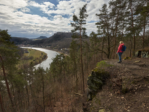 Johann Alexander Thiele View A person in a red jacket and blue rucksack stands on a rocky outcrop and looks out over a river in a wooded landscape under a cloudy sky.