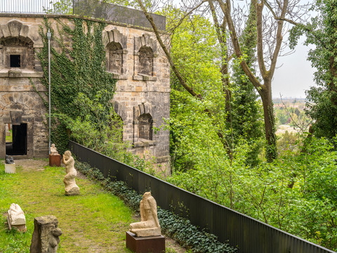 Bastions van kasteel Sonnenstein Alte Steinmauer des Schloss Sonnenstein mit Efeu bewachsen, umgeben von Bäumen; im Vordergrund stehen mehrere antike Skulpturen auf einer grünen Wiese.